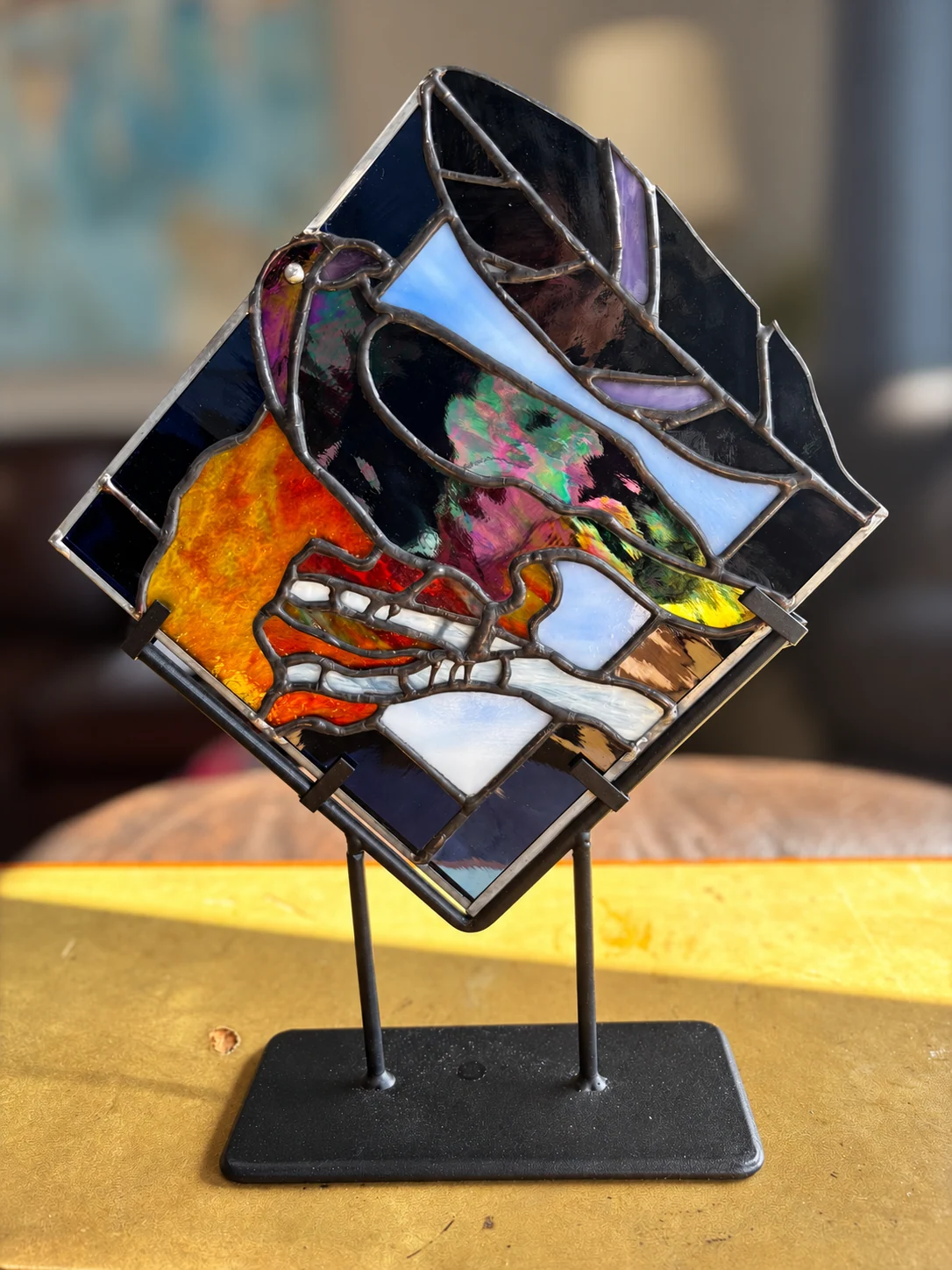 Crow stained-glass panel on its tabletop stand, with pink and green iridescence and a softly blurred room behind it